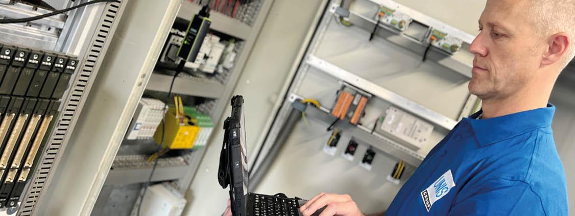 Technician using a laptop while checking industrial control equipment inside an electrical cabinet.